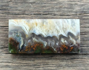 May include: A rectangular stone with a landscape design. The stone features a mix of colours, including white, beige, brown, green, and orange. The design resembles a natural landscape with layers of colour and texture. The stone is set against a wooden background.