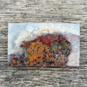 Peut inclure: Une pierre rectangulaire polie avec un motif de paysage. La pierre présente un mélange de couleurs, dont le blanc, l'orange, le rouge et le vert, ressemblant à une scène naturelle. La pierre est posée sur un fond en bois.