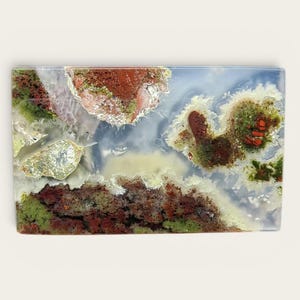 Op de afbeelding: Rechthoekige steen met een landschapsontwerp. De steen heeft een mix van kleuren, waaronder rood, groen en wit, met een doorschijnend uiterlijk. Het ontwerp lijkt op een natuurlijk landschap.