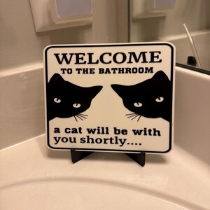 May include: A white sign with black text that reads "WELCOME TO THE BATHROOM" and "a cat will be with you shortly...." Two black cat silhouettes are on the sign. The sign is on a black stand.