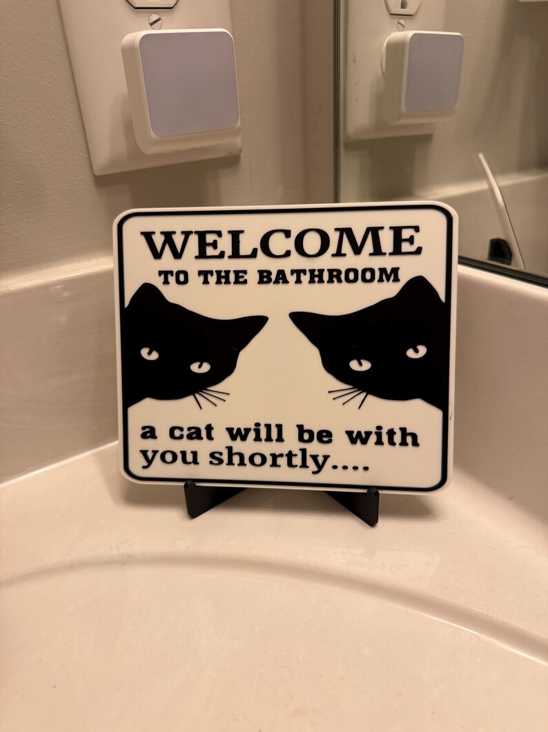 May include: A white sign with black text that reads "WELCOME TO THE BATHROOM" and "a cat will be with you shortly...." Two black cat silhouettes are on the sign. The sign is on a black stand.