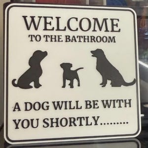 May include: White square sign with black text and dog silhouettes. The sign reads "WELCOME TO THE BATHROOM" and "A DOG WILL BE WITH YOU SHORTLY..." Three black dog silhouettes are featured on the sign.