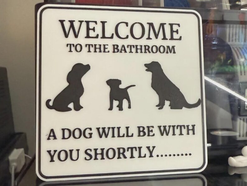 May include: White square sign with black text and dog silhouettes. The sign reads "WELCOME TO THE BATHROOM" and "A DOG WILL BE WITH YOU SHORTLY..." Three black dog silhouettes are featured on the sign.