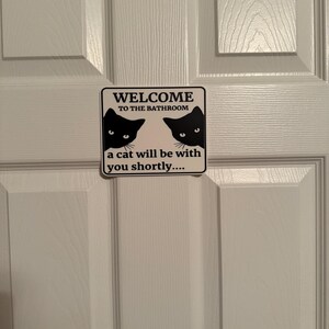 May include: A rectangular sign with the text "WELCOME TO THE BATHROOM" and "a cat will be with you shortly...." is affixed to a white door. The sign features two black cat silhouettes.