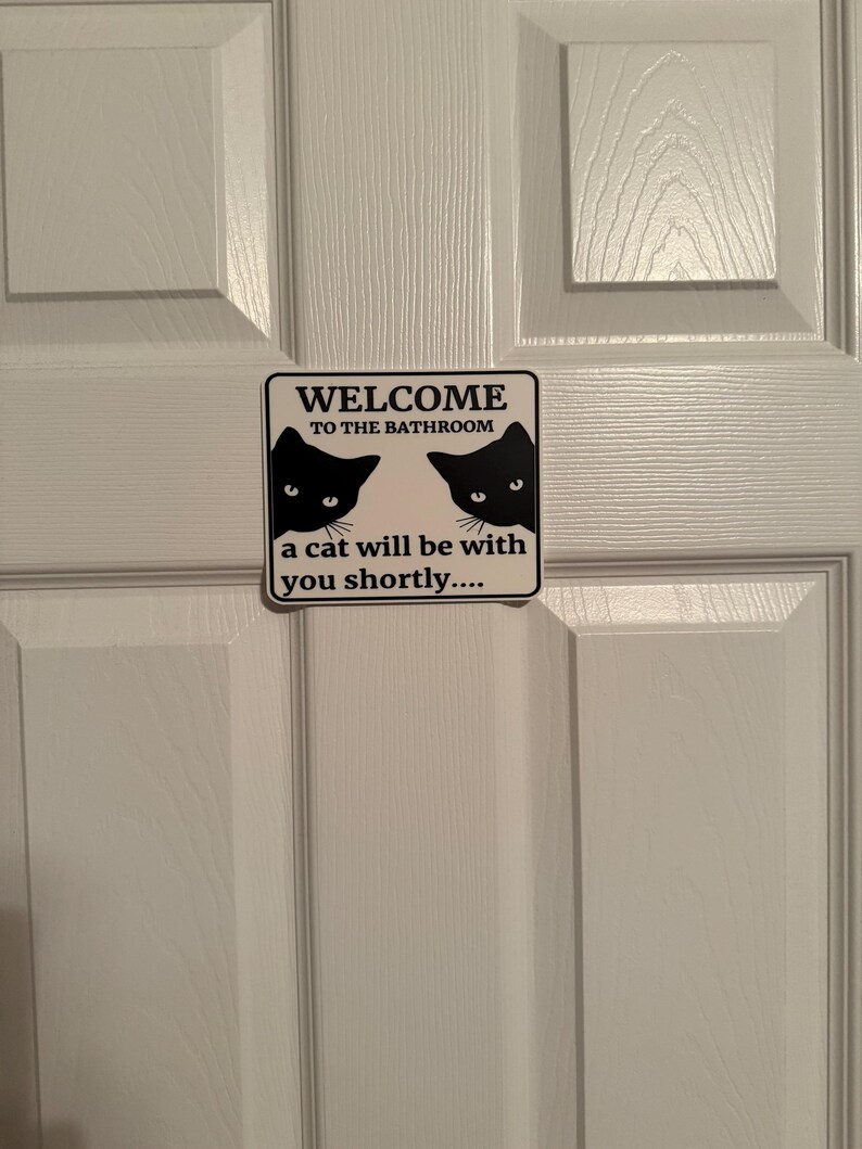 May include: A rectangular sign with the text "WELCOME TO THE BATHROOM" and "a cat will be with you shortly...." is affixed to a white door. The sign features two black cat silhouettes.