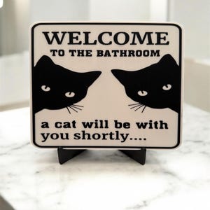 May include: A rectangular sign with the text "WELCOME TO THE BATHROOM" and "a cat will be with you shortly...." in black. Two black cat silhouettes are on the sign. The sign is on a black stand.