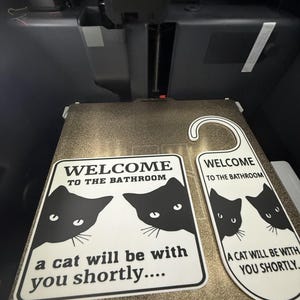 May include: Two white bathroom signs with black cat graphics and the text "WELCOME TO THE BATHROOM" and "A CAT WILL BE WITH YOU SHORTLY." One sign is rectangular, the other is a door hanger shape.
