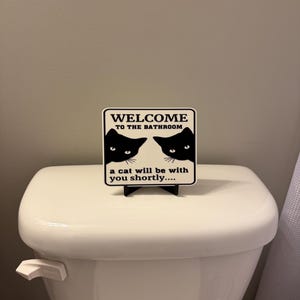 May include: A square sign with the text "WELCOME TO THE BATHROOM" and two black cat silhouettes. Below the text, it reads "a cat will be with you shortly...." The sign is placed on top of a white toilet.