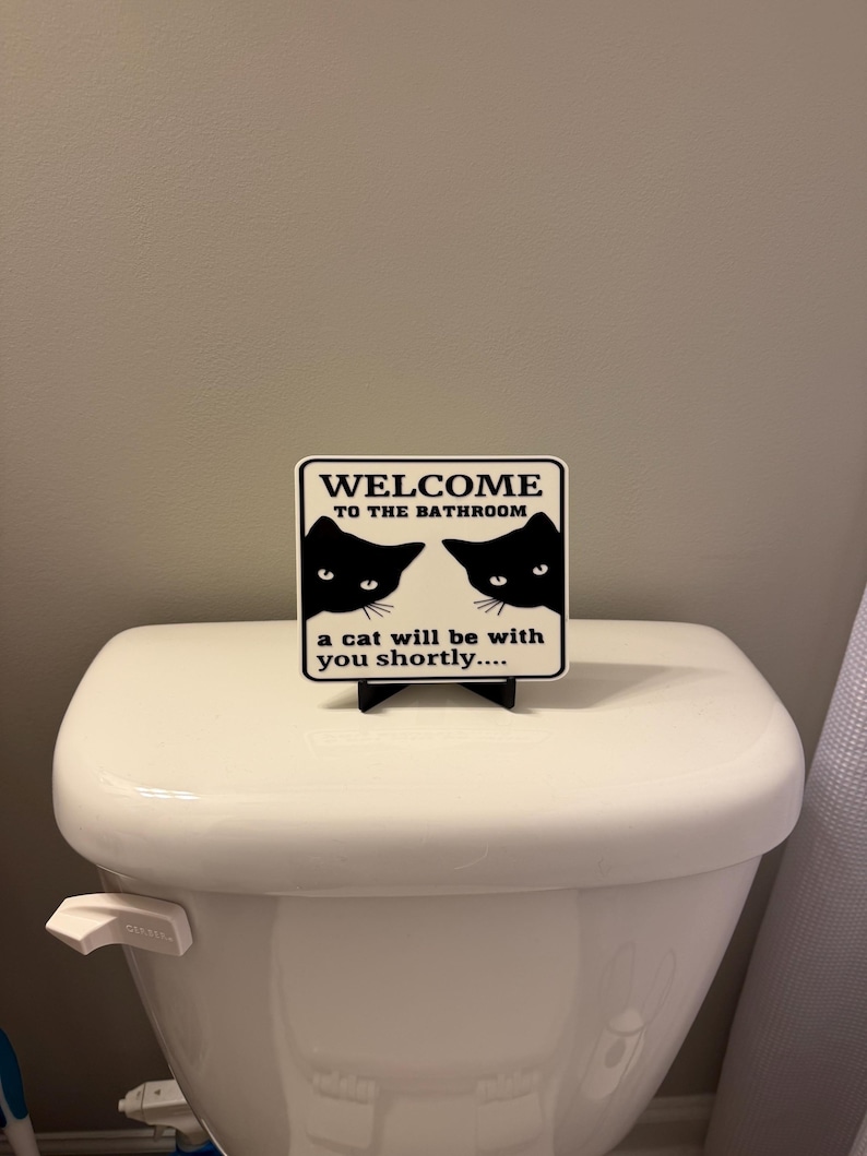 May include: A square sign with the text "WELCOME TO THE BATHROOM" and two black cat silhouettes. Below the text, it reads "a cat will be with you shortly...." The sign is placed on top of a white toilet.