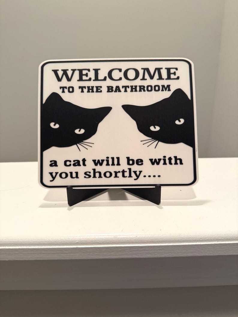 May include: A white sign with black text and two black cat silhouettes. The sign reads "WELCOME TO THE BATHROOM" and "a cat will be with you shortly...." The sign is displayed on a black stand.