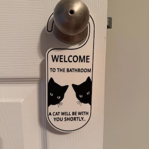 May include: White door hanger with the text "WELCOME TO THE BATHROOM" and "A CAT WILL BE WITH YOU SHORTLY..". The design includes two black cat silhouettes peeking from the sides.