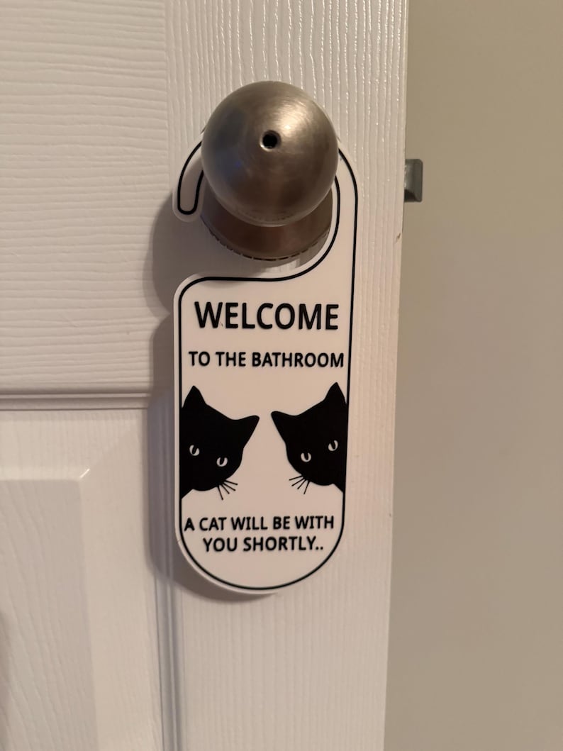 May include: White door hanger with the text "WELCOME TO THE BATHROOM" and "A CAT WILL BE WITH YOU SHORTLY..". The design includes two black cat silhouettes peeking from the sides.