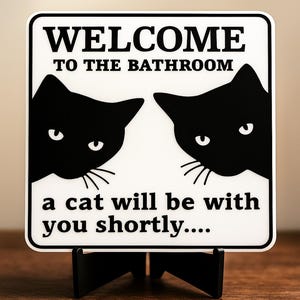 May include: A white sign with black text that reads "WELCOME TO THE BATHROOM" and "a cat will be with you shortly...." Two black cat silhouettes are on the sign. The sign is on a black stand.