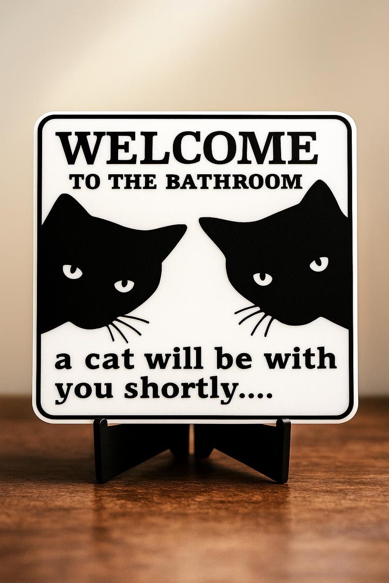 May include: A white sign with black text that reads "WELCOME TO THE BATHROOM" and "a cat will be with you shortly...." Two black cat silhouettes are on the sign. The sign is on a black stand.