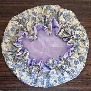 Könnte beinhalten: Eine Duschhaube mit floralem Muster und einem hellvioletten Satinfutter und -besatz. Die Haube hat einen cremefarbenen Hintergrund mit blauen und violetten Blumenmustern. Die Innenseite ist einfarbig hellviolett.