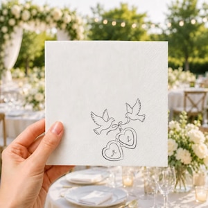 May include: A square white paper napkin featuring a black line art illustration of two flying doves holding a pair of heart-shaped lockets. The left locket contains the letter M, and the right locket contains the letter L, both in a cursive font.