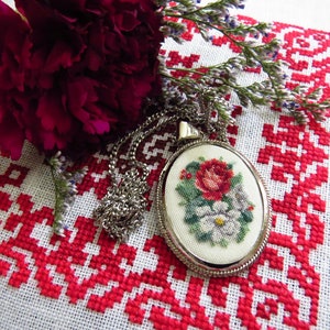 Könnte beinhalten: Eine silberne ovale Halskette mit einem Kreuzstich-Design einer roten Rose und weißen Blumen auf weißem Hintergrund. Die Halskette liegt auf einem weißen Stoff mit einem roten Kreuzstich-Muster.