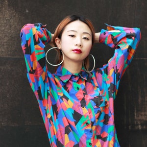 May include: A button-down shirt with a vibrant geometric pattern in blue, red, green, and black. The shirt is paired with blue jeans. Large silver hoop earrings complete the look.