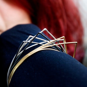 May include: A contemporary bracelet crafted from silver, black, and gold wire. The design incorporates a series of geometric shapes, offering a modern, minimalist aesthetic. The bracelet is displayed on a dark blue sleeve, with a blurred background.