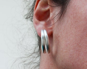 May include: Silver double hoop earrings. These earrings have a smooth, polished finish and a modern, minimalist design. The earrings are worn on the ear and are a bright, shiny silver color. The earrings are a simple yet elegant accessory.
