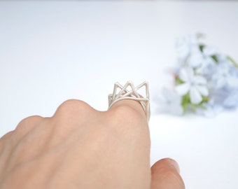 May include: A silver geometric ring with a unique angular design. The ring is worn on a finger, showcasing its intricate, openwork structure. The ring's design features multiple triangular shapes, creating a modern and artistic look.