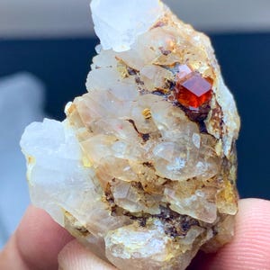 May include: A natural mineral specimen featuring a cluster of clear quartz crystals and a vibrant, reddish-orange garnet crystal. The quartz crystals have a crystalline structure, and the garnet is a well-formed, geometric shape. The overall color palette includes clear, white, and orange hues.
