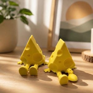 May include: Two whimsical yellow cheese wedge figurines with holes and attached legs are displayed on a wooden surface. The cheese wedges are in a relaxed pose, suggesting a playful design. A plant in a white pot and a framed picture are in the background.