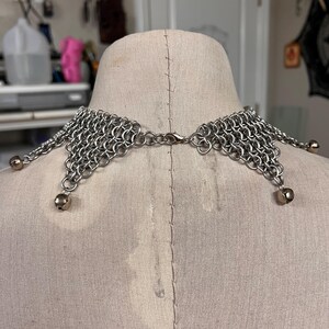 Puede incluir: Un collar de cota de malla plateada con un dise&ntilde;o decorativo. El collar presenta una serie de peque&ntilde;os anillos de metal entrelazados, formando un cuello flexible y drapeado. Peque&ntilde;as cuentas redondas plateadas cuelgan de los bordes inferiores.