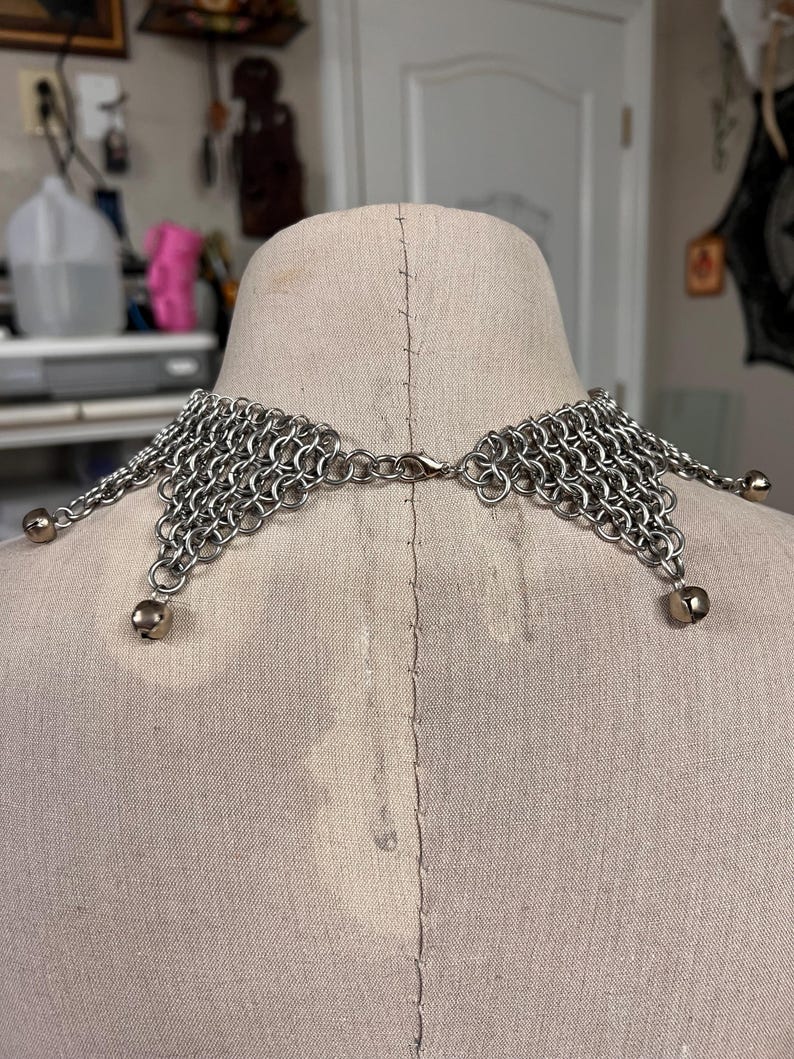 Puede incluir: Un collar de cota de malla plateada con un dise&ntilde;o decorativo. El collar presenta una serie de peque&ntilde;os anillos de metal entrelazados, formando un cuello flexible y drapeado. Peque&ntilde;as cuentas redondas plateadas cuelgan de los bordes inferiores.