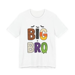 May include: A white short-sleeved t-shirt featuring the text BIG BRO in a Halloween-themed font. The letters are decorated with orange and black stripes, black polka dots, candy corn colors, mummy bandages, purple, and green slime patterns.