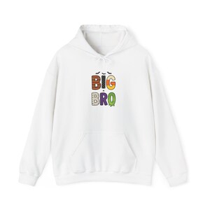 Pode incluir: Um moletom com capuz branco apresentando um gráfico no peito com o texto BIG BRO. As letras são decoradas com vários padrões, incluindo listras laranjas e pretas, pontos brancos, milho doce, faixas de múmia e lodo verde escorrendo.