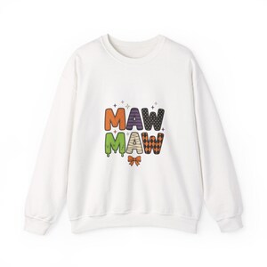 May include: A white long-sleeve sweatshirt featuring a graphic print of the text MAW MAW in colorful, patterned letters. The letters include orange, purple stripes, black polka dots, green slime, mummy bandages, and orange diamonds, with a small bow.