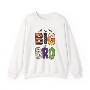 May include: A white long-sleeve sweatshirt featuring the text BIG BRO in colourful, patterned letters. The letters include orange and black stripes, black polka dots, candy corn, mummy bandages, purple, and green slime. Three bats and a spider are included.