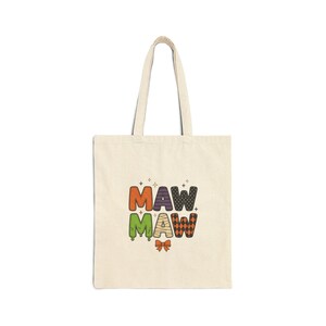 May include: A beige canvas tote bag featuring the text MAW MAW in a multi-colored Halloween-themed font. The letters are decorated with orange, purple, green, mummy-wrap, and black diamond patterns, accented by a small orange bow at the bottom.