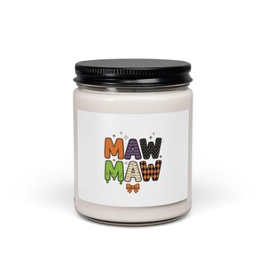 May include: A glass candle jar with a black lid and a white label featuring the text MAW MAW in colorful, patterned letters. The letters include orange, purple stripes, black polka dots, green slime, mummy bandages, and orange diamonds with a bow.