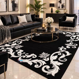 May include: A rectangular area rug featuring a solid black background with a prominent white and light gray scrolling acanthus leaf border pattern. The design includes a small logo in the corner and covers a large portion of the floor space.