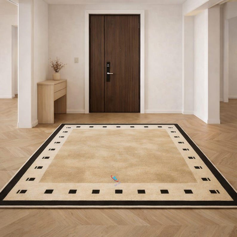 Pode incluir: Um tapete retangular bege com uma borda preta e detalhes quadrados. O tapete est&aacute; colocado sobre um ch&atilde;o de madeira em um quarto bem iluminado, com uma porta marrom escura ao fundo. O design do tapete adiciona um toque de eleg&acirc;ncia ao espa&ccedil;o.
