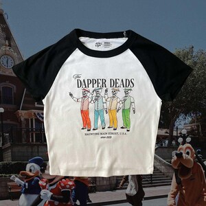 May include: White and black baseball tee shirt with a colorful graphic of four skeletons in suits with the text "The Dapper Deads Haunting Main Street, U.S.A. since 1959"