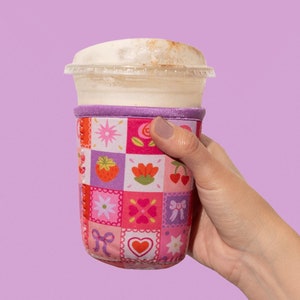 Pode incluir: Um protetor de copo estampado em rosa e roxo com um copo branco cheio de uma bebida marrom clara. O protetor de copo tem uma variedade de pequenas flores rosa, vermelhas e roxas, corações e morangos.