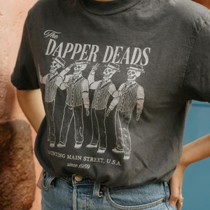 May include: Dark gray t-shirt with the text "The Dapper Deads" and an illustration of four skeleton figures in suits and hats. The text "Haunting Main Street, U.S.A. since 1959" is printed below the figures. The shirt is paired with blue jeans.