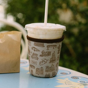 May include: A brown paper bag sits next to a white cup with a brown patterned sleeve. The cup has a straw sticking out of it and is filled with a light brown liquid. The cup and bag are on a blue table with a white floral design.
