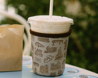 May include: A brown paper bag sits next to a white cup with a brown patterned sleeve. The cup has a straw sticking out of it and is filled with a light brown liquid. The cup and bag are on a blue table with a white floral design.