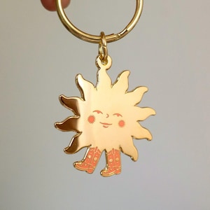 May include: Gold-colored keychain featuring a smiling sun with orange boots. The sun has a yellow face with red cheeks and a small smile.