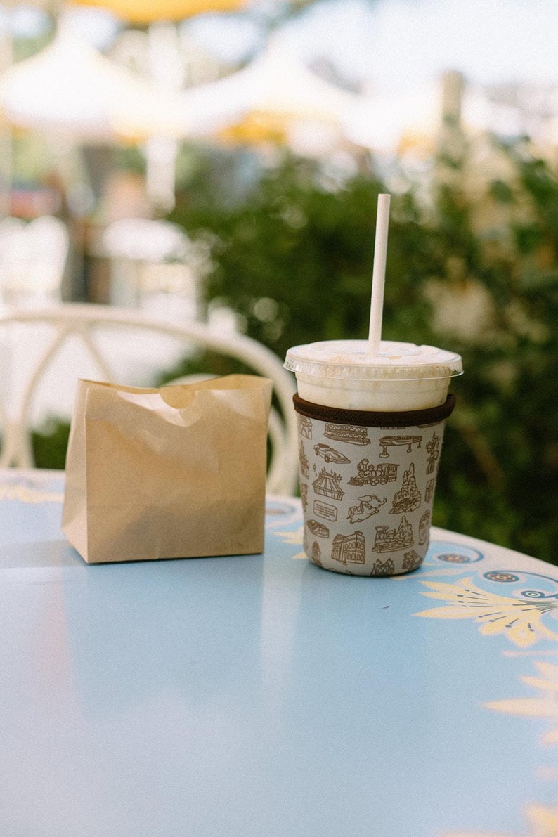 May include: A brown paper bag sits next to a white cup with a brown patterned sleeve. The cup has a straw sticking out of it and is filled with a light brown liquid. The cup and bag are on a blue table with a white floral design.