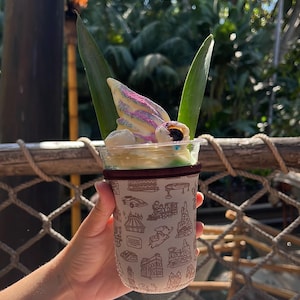May include: A white cup with a green and brown patterned sleeve, holding a pineapple Dole Whip with a swirl of purple and white soft serve ice cream topped with a pineapple leaf and a chocolate candy eye.
