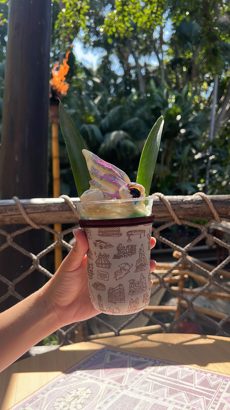 May include: A white cup with a green and brown patterned sleeve, holding a pineapple Dole Whip with a swirl of purple and white soft serve ice cream topped with a pineapple leaf and a chocolate candy eye.