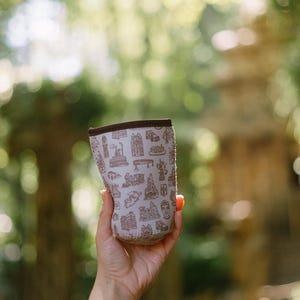 May include: A white neoprene can cooler with a brown print of various buildings and landmarks. The cooler is being held in a person's hand.