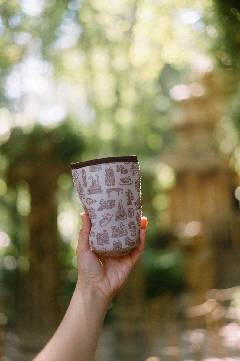 May include: A white neoprene can cooler with a brown print of various buildings and landmarks. The cooler is being held in a person's hand.