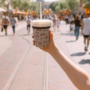 May include: A white to-go cup with a brown sleeve featuring a pattern of various Disney park attractions. The cup is filled with a light brown beverage and has a clear plastic lid with a straw.