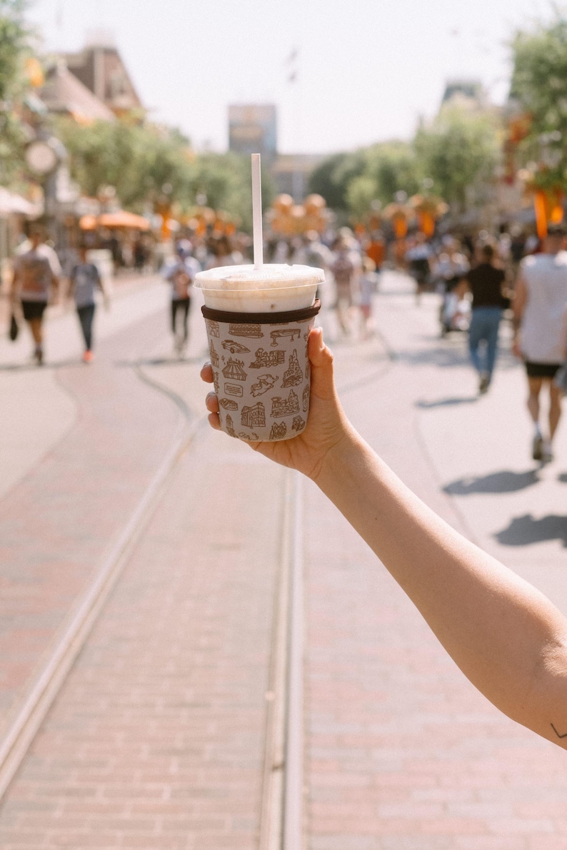 May include: A white to-go cup with a brown sleeve featuring a pattern of various Disney park attractions. The cup is filled with a light brown beverage and has a clear plastic lid with a straw.